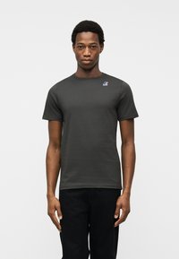 Νεαρός άνδρας που φοράει σκούρο γκρι κοντομάνικο μπλουζάκι με μικρό πολύχρωμο λογότυπο στο αριστερό μέρος του στήθους, στέκεται μπροστά σε απλό ανοιχτό φόντο.