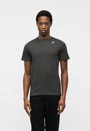 Giovane uomo che indossa una T-shirt a maniche corte di colore grigio scuro con un piccolo logo multicolore sul petto a sinistra, in piedi davanti a uno sfondo chiaro e uniforme.