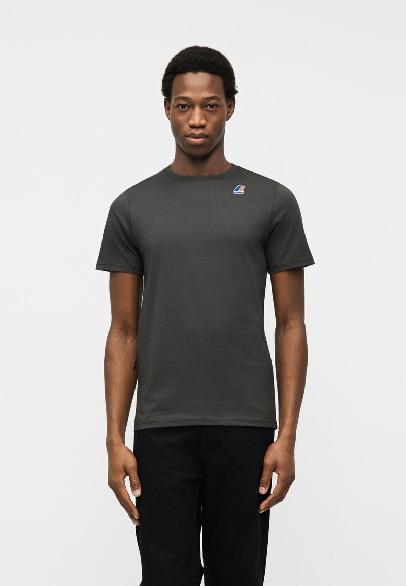 Νεαρός άνδρας που φοράει σκούρο γκρι κοντομάνικο μπλουζάκι με μικρό πολύχρωμο λογότυπο στο αριστερό μέρος του στήθους, στέκεται μπροστά σε απλό ανοιχτό φόντο.