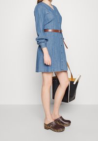 Jeansowa sukienka z długimi rękawami, zapinana na guziki, z brązowym paskiem; w połączeniu z brązowymi chodakami z złotymi ćwiekami; trzyma czarną torbę na ramię.