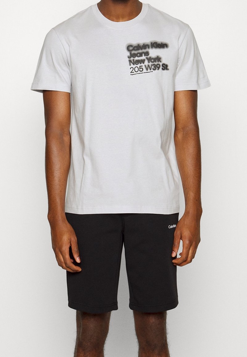 Bílá bavlněná T-shirt s černým grafickým textem "Calvin Klein Jeans New York 205 W39 St." a černé sportovní šortky s logem.