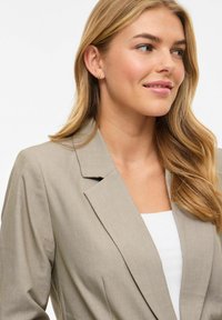 Femme souriante aux longs cheveux blonds, portant un blazer beige, un haut blanc et de petites boucles d'oreilles en or, regardant vers la droite sur fond blanc.