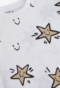Camiseta de algodón blanca con estrellas sonrientes marrones dibujadas a mano y caras sonrientes negras en la tela. Textura suave, diseño de cuello redondo.