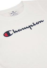 Camiseta de algodón blanca con un logo "Champion" en azul marino y un acento rojo en el lado izquierdo. Escote redondeado y mangas cortas.