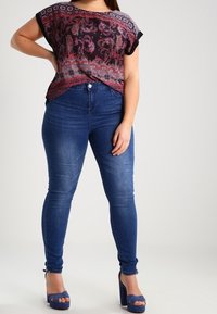 Kortærmet mønstreret top i sort, rød og lilla, parret med slim-fit blå jeans. Modellen har blå højhælede sko på.