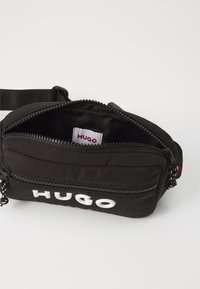 Sac en tissu noir avec fermeture éclair, comportant un logo blanc "HUGO" sur le devant et une surface texturée. Étiquette intérieure visible.