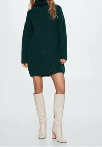 Robe pull tricotée vert foncé avec col roulé, manches longues et fentes latérales, associée à des bottes beiges à talons montant jusqu'aux genoux.