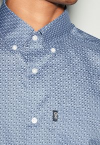 Een blauwe overhemden met een gestructureerd patroon. Voorzien van witte knopen en een klein zwart label met een logo op de linkerborst.