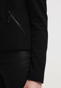 Černá bunda se zipovou kapsou a texturovaným materiálem. Přiléhavý střih, viditelné švy a elegantní silueta doplněná černými kalhotami.