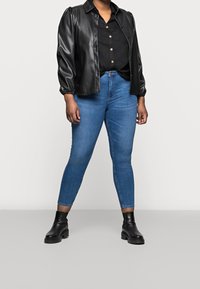 Persona que lleva una camisa negra de botones, una chaqueta de cuero negra, unos vaqueros ajustados azules y botas negras hasta el tobillo, de pie sobre un suelo gris liso.