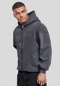 Sweat à capuche gris zippé avec une grande capuche, des poignets élastiques et un logo discret sur la poitrine. Tissu doux et texturé avec une coupe décontractée.