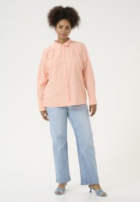Camisa de botón de manga larga color rosa claro con cuello, de corte relajado. Combinada con jeans rectos de tiro alto en azul claro y tacones abiertos.