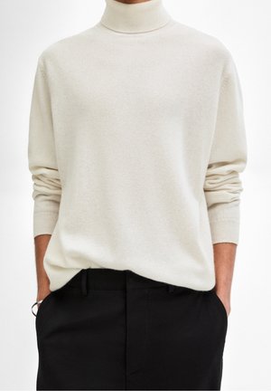 Personne portant un pull à col roulé crème à manches longues et un pantalon noir, mains dans les poches, fond neutre.