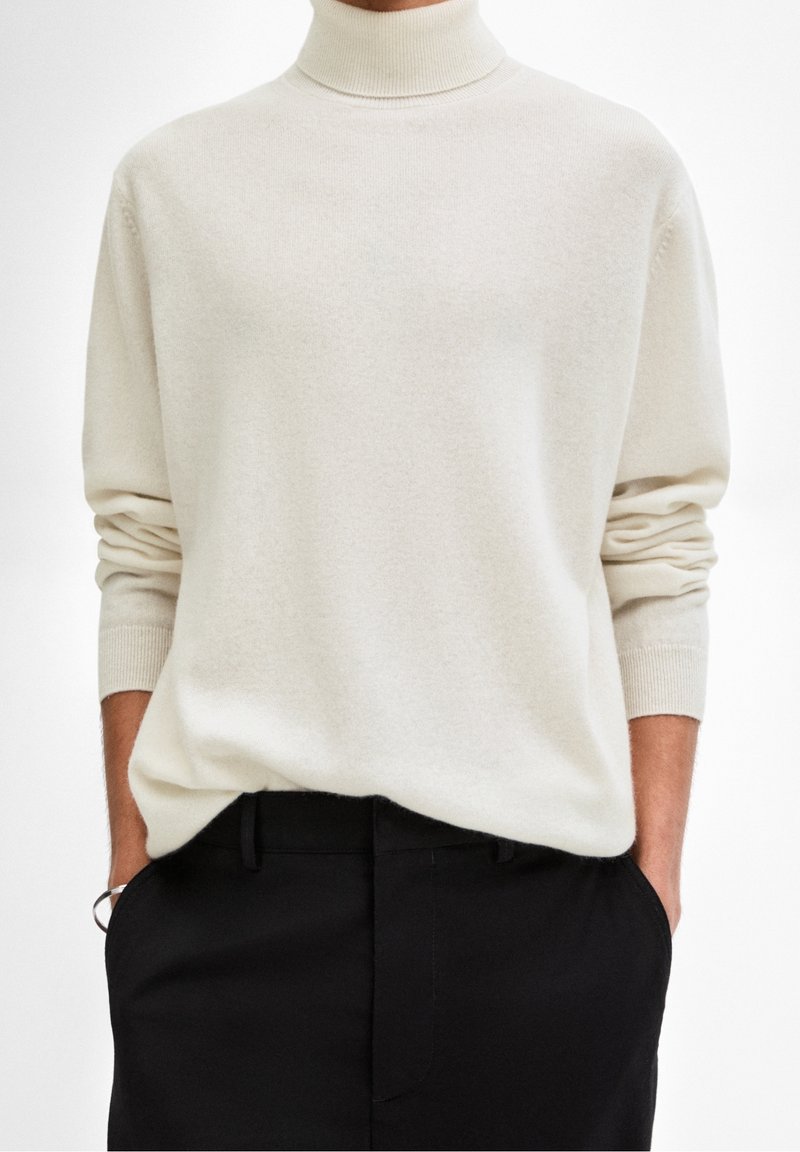 Personne portant un pull à col roulé crème à manches longues et un pantalon noir, mains dans les poches, fond neutre.