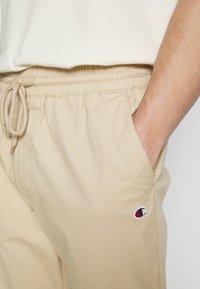 Béžové šortky z bavlněné směsi mají elastický pás s tkaničkou, boční kapsy a malou vyšitou nášivku blízko lemu.