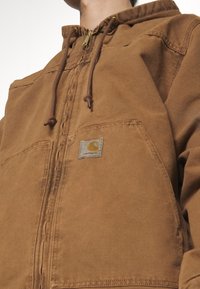 Brun zip-up hættejakke lavet af slidstærkt materiale. Har en forlommes og snoretræk; inkluderer et logo-patch på venstre side.