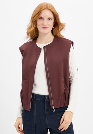 Bordeaux mouwloze vest gemaakt van een gladde stof, met een rits aan de voorkant, zijzakken en een relaxte pasvorm. Draagt over een wit langemouwen top.