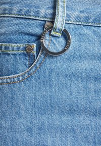 Světle modré džíny z denimu mají kruhový kovový kroužek připevněný k poutku na opasek, doplněný kontrastním žlutým prošitím a malým emblémem na kapse.