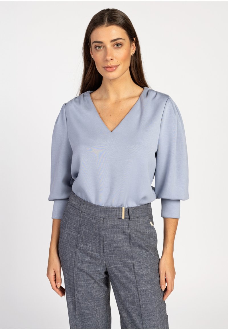 Lichtblauwe blouse met een V-hals, puffmouwen en een gladde textuur, gecombineerd met grijs geruite broek met een platte voorkant en gouden accenten.