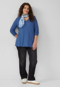 Blauwe langemouwen V-hals t-shirt met een relaxte pasvorm, gecombineerd met zwarte jeans. Een lichtblauwe met patroon sjaal voegt kleur en textuur toe. Witte sneakers.