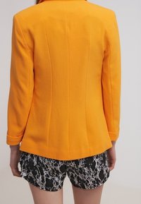 Orange blazer with a structured fit, featuring seam detailing. Paired with black and white marble-patterned shorts.