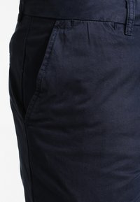 Mørkeblå bomuldsbukser med en glat tekstur, udstyret med en sidelomme og en skræddersyet pasform. Stoffet er let reflekterende under lys.