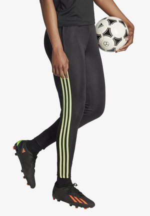 Osoba ubrana w czarne spodnie sportowe z jasnozielonymi paskami i czarne korki trzymająca czarno-białą piłkę nożną Adidas.