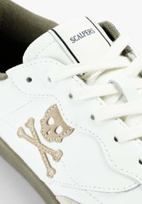Zapatilla blanca con bordado de calavera y huesos en beige, cordones blancos y texto "Scalpers" en la lengüeta.