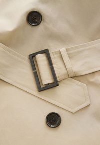 Tela de gabardina beige con una hebilla rectangular negra y un botón negro redondo con un diseño floral. Textura suave con costuras visibles.