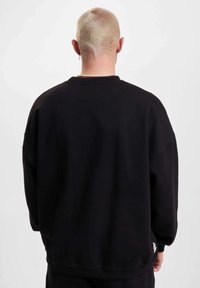 Mustat ylipitkät hupparit, joissa on ribbineuletettu pyöreä kaula-aukko, laskeutuvat olkapäät ja pieni valkoinen logo helman lähellä. Pehmeä, teksturoidusta kankaasta valmistettu.