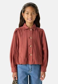 Veste couleur rouille avec des manches longues bouffantes, une fermeture à boutons et un col. Portée sur un jean bleu clair, avec un bandeau à imprimé léopard.