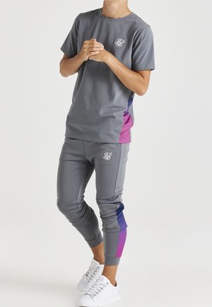 Persona con un conjunto deportivo gris con paneles laterales morados y azules, zapatillas blancas, de pie con las manos entrelazadas sobre un fondo blanco liso.