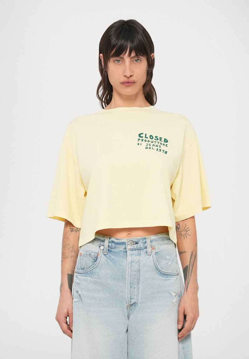 T-shirt jaune raccourci à manches courtes, avec un texte graphique vert sur la poitrine. Associé à un jean bleu clair déchiré.