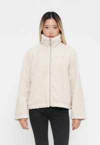 Veste zippée beige clair, floue, avec un col montant et des manches longues et larges. Dotée d'un tissu texturé. Associée à un pantalon noir.