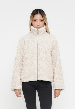 Ljus beige, fluffig zip-up jacka med hög krage och långa, vida ärmar. Har en strukturerad tyg. Stylad med svarta byxor.