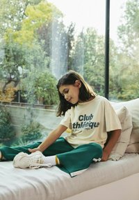Camiseta crema con "Club athlétique" en azul marino, combinada con pantalones anchos verdes. Zapatillas blancas con acentos amarillos, sentadas sobre un cojín gris.