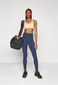 Sostenedor deportivo beige, leggings navy de cintura alta, zapatos negros y un bolso negro con correa. La modelo se encuentra de pie contra un fondo liso.