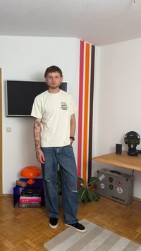 T-shirt de algodão creme com gráfico verde, jeans de denim de corte largo com detalhes desgastados e sapatos slip-on pretos. O ambiente apresenta listras coloridas nas paredes.