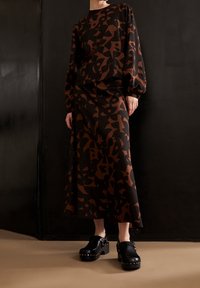 Nainen, jolla on yllään pitkähihainen musta ja ruskea abstraktikuosinen mekko ja mustat niititetyt puukengät, seisoo tumman seinän edessä.