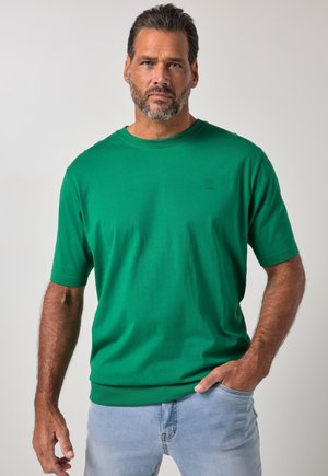 Homme avec une barbe et des cheveux courts portant une chemise verte à manches courtes et un jean bleu clair, debout avec une main dans la poche.