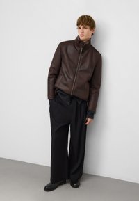 Bruine leren jas met een hoge kraag, ritssluiting aan de voorkant en minimale stiksels. Gecombineerd met een donkergrijze trui en losse zwarte broek.