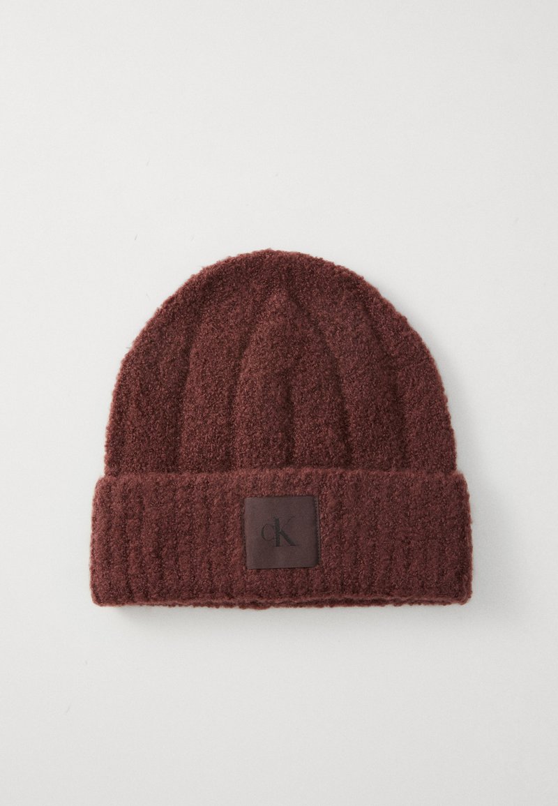 Bonnet tricoté bordeaux en tissu doux et texturé avec un design à côtes. Il est orné d'un patch en cuir marron avec le logo "ck" à l'avant.