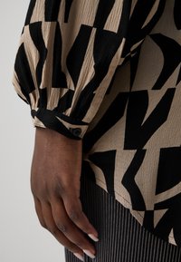 Haut beige à manches longues avec des motifs abstraits noirs. Tissu texturé, poignets ajustés avec un détail de bouton. Ondulations et drapés subtils.