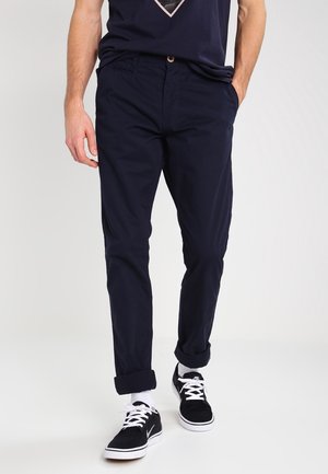 Mand iført navy blå afslappede bukser med opsmøgede bukseben, sorte og hvide sneakers, stående med den ene hånd i lommen på en ensfarvet baggrund.