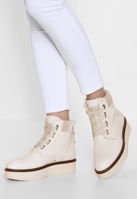 Jasno beżowe botki za kostkę z teksturowanym wykończeniem, z sznurowanym zamknięciem i kontrastującą grubą podeszwą. Z boku zamek błyskawiczny ułatwiający zakładanie.