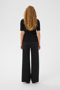 Osoba s dlhými kučeravými vlasmi, oblečená v čiernom krátkom rukáve s rebrovaním a širokých čiernych nohaviciach, stojaca pred jednostranným pozadím.