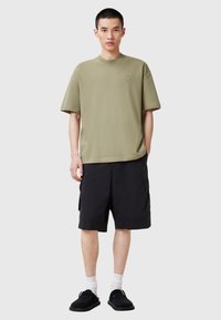 Una t-shirt verde oliva a maniche corte con scollo rotondo e dettaglio del logo; abbinata a pantaloni cargo neri e scarpe slip-on nere.