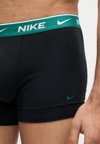 Άνδρας φοράει μαύρο Nike boxer σλιπ με πράσινη μέση και μικρό πράσινο λογότυπο Nike στο αριστερό μηρό.