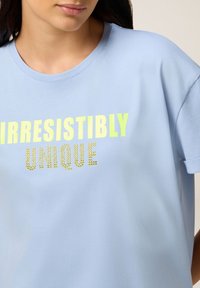 Camiseta de manga corta de color azul claro con el texto llamativo en amarillo "IRRESISTIBLEMENTE ÚNICO" adornado con piedras brillantes.