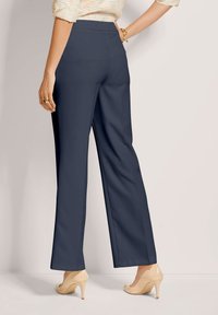 Vrouw in marineblauwe wijde broek en beige hoge hakken, met één hand op de heup en een beige textuurtop die zichtbaar is bij de taille.